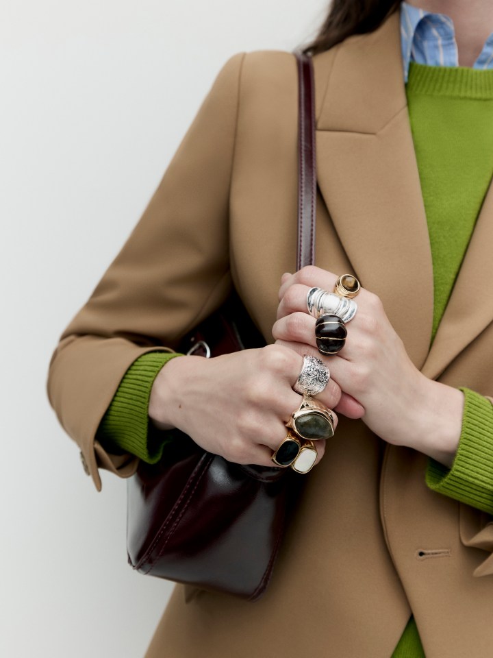 Hands with multiple rings holding a dark red handbag.