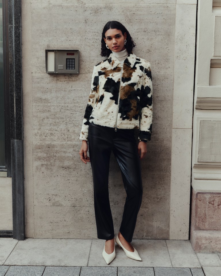 A woman modeling the M&S Autumn Winter 2025 collection wearing a white turtleneck, a faux fur jacket with a black, white, and brown cow print, black leather pants, and white pointed-toe heels.