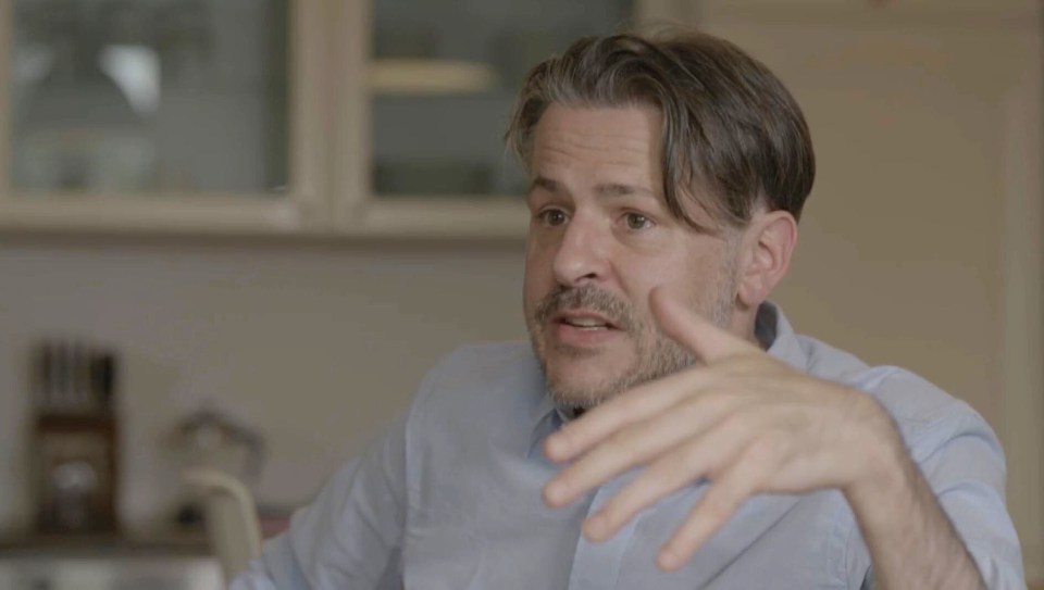 A man with graying hair and a beard, wearing a light blue shirt, gestures with his right hand.