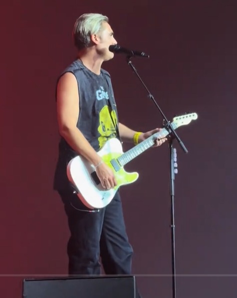 Man with grey hair in a sleeveless t-shirt playing a white and yellow electric guitar and singing into a microphone on stage.