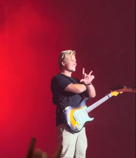 A musician on stage with a guitar, making a rock and roll hand gesture, under a red light with text "Busted discussing James Bourne's absence and announcing his replacement for the tour".