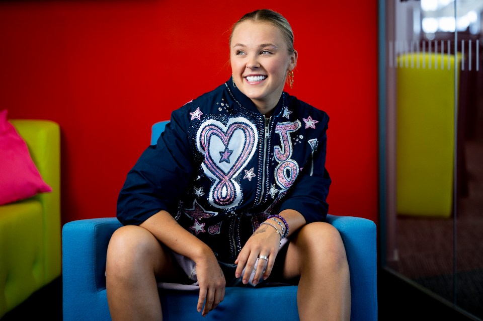 JoJo Siwa sitting in a blue chair, smiling and looking to the right.