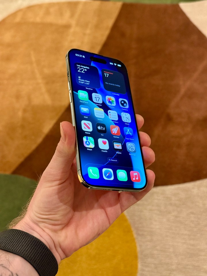 A hand holding an iPhone displaying the home screen with various app icons, time, weather, and calendar widgets.