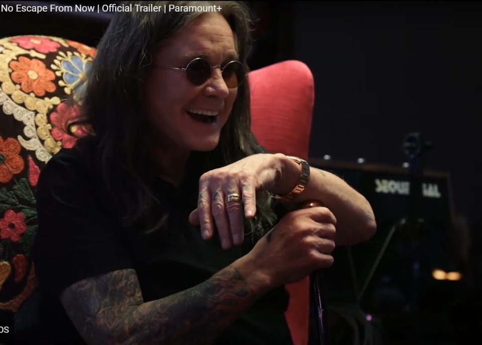 Ozzy Osbourne in dark sunglasses, a black shirt, and tattooed arms, holding a cane, laughing in a chair.