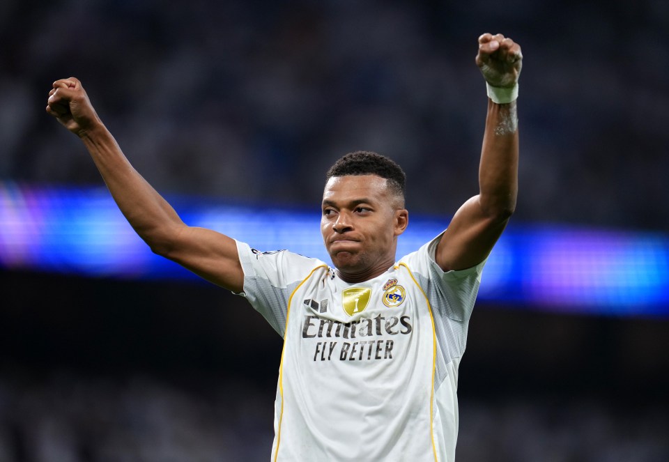 Kylian Mbappe in a Real Madrid jersey celebrates scoring a goal, arms raised.