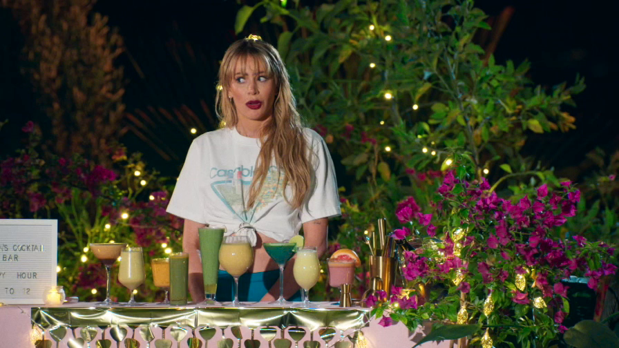 A woman with blonde hair, wearing a white t-shirt, stands behind a bar with several colorful cocktails.