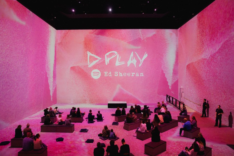 Audience members sitting on benches and cushions in a room with pink lighting and "PLAY Ed Sheeran" projected on the back wall.