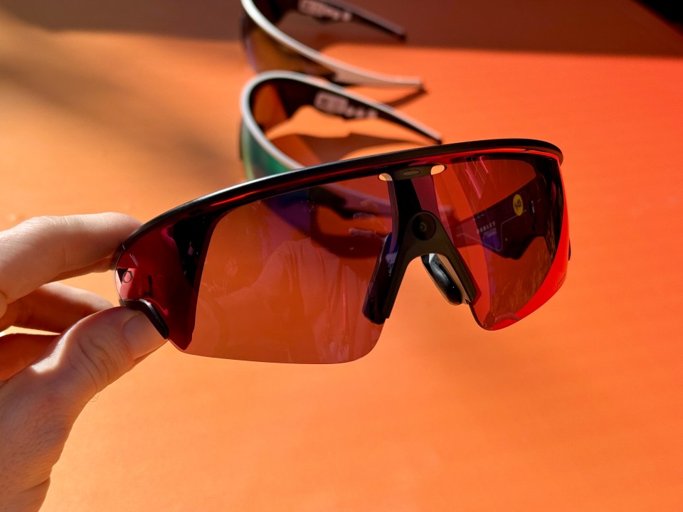 A hand holding a pair of sports sunglasses with orange lenses, with two other similar pairs blurred in the background on an orange surface.