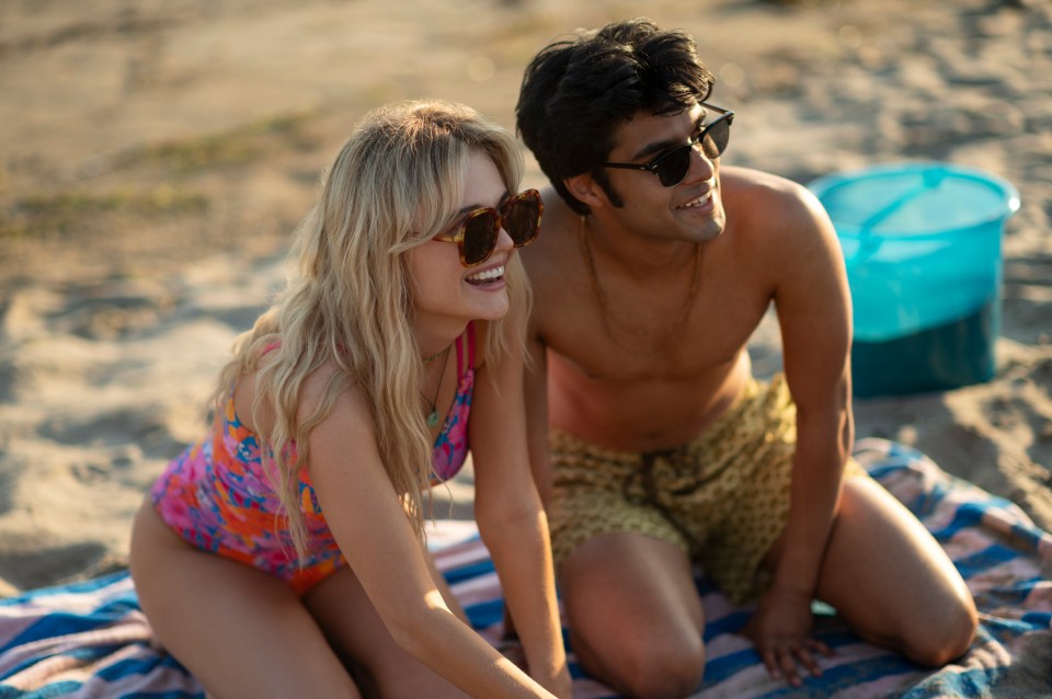 Emily Alyn Lind as Cadence Sinclair and Shubham Maheshwari as Gat sit on a striped towel on a sandy beach.