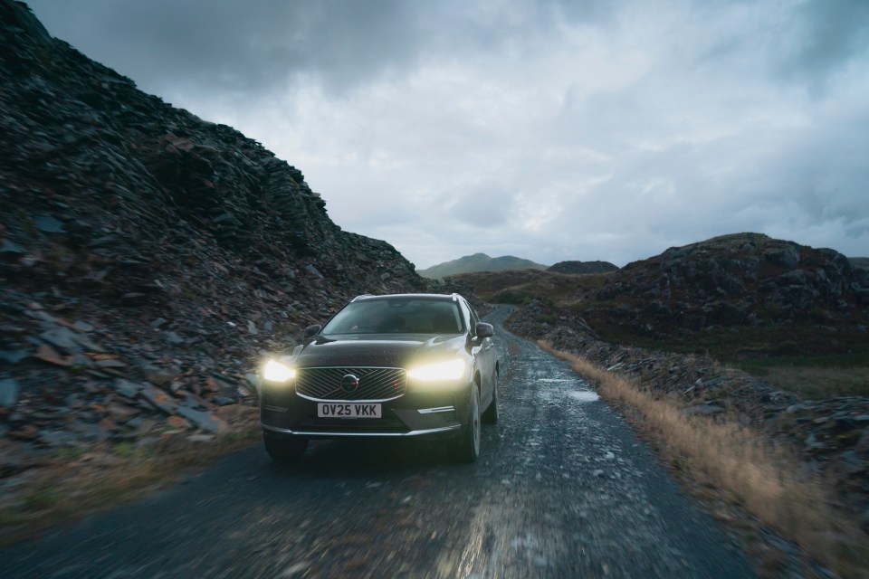 A dark Volvo XC60 driving on a wet, rocky road through a mountainous landscape under a cloudy sky.