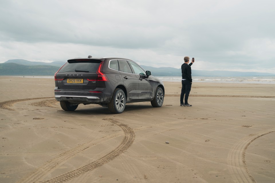A man standing on a sandy beach next to a Volvo XC60 and taking a photo of the ocean.