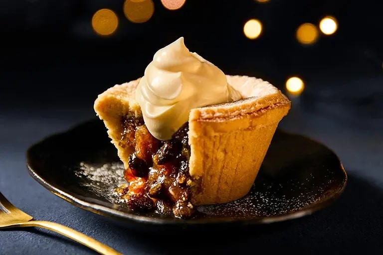 A mince pie with a dollop of cream on top, against a dark background with blurred lights.