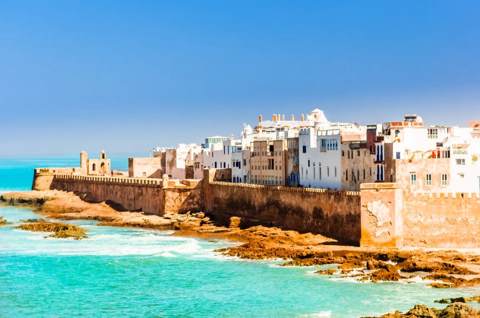 Aerial view of the old city of Essaouira with white buildings behind a stone fortress wall, next to turquoise ocean water and brown rocks.