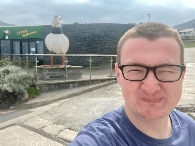 Rich James Cross, 27, in a blue shirt and glasses, smiling for a selfie in front of a building with a "Croeso Welcome" sign and a large bird statue.
