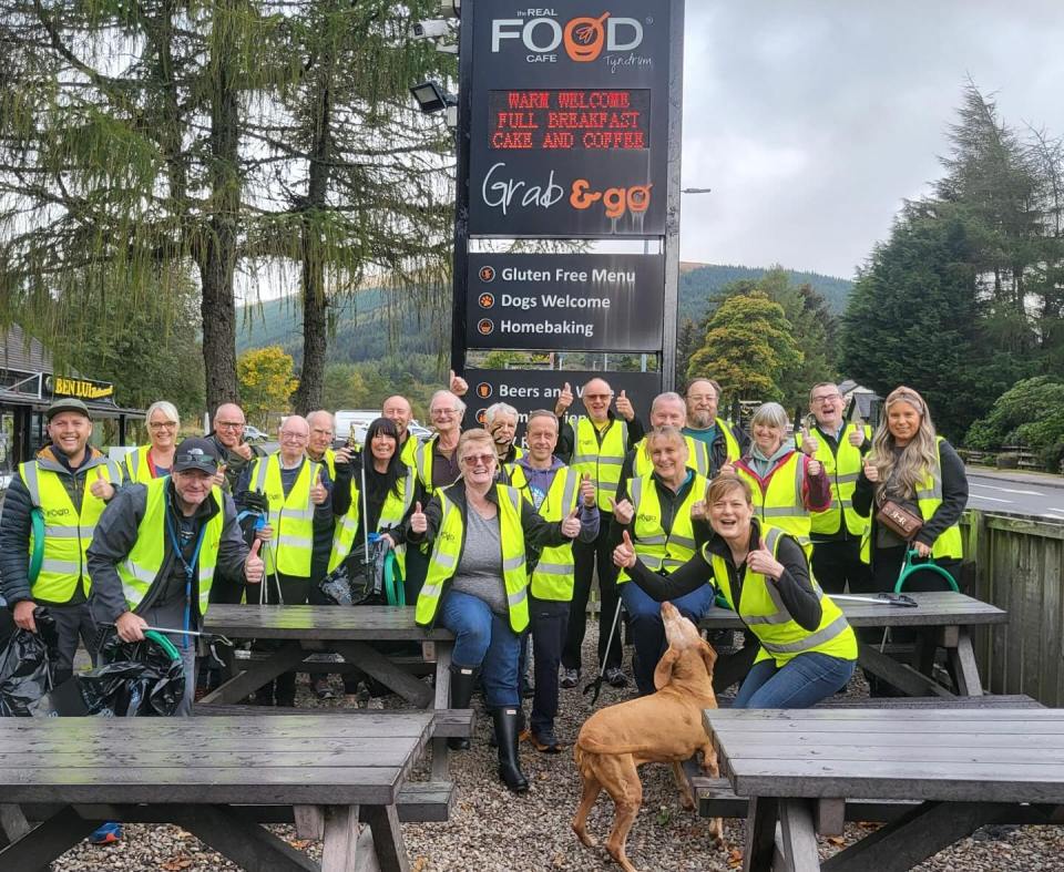 A group of people wearing bright yellow vests and giving thumbs up outside "The Real Food Cafe" in Tyndrum, with a dog by a picnic table.