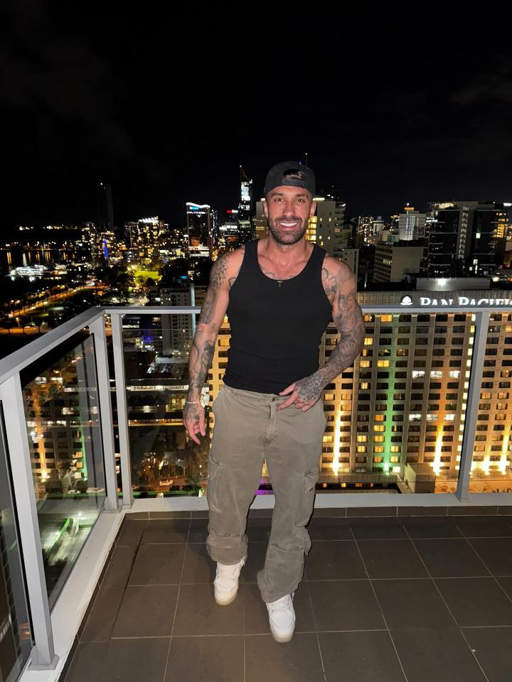 Billy Belcher standing on a balcony at night with a city skyline in the background.