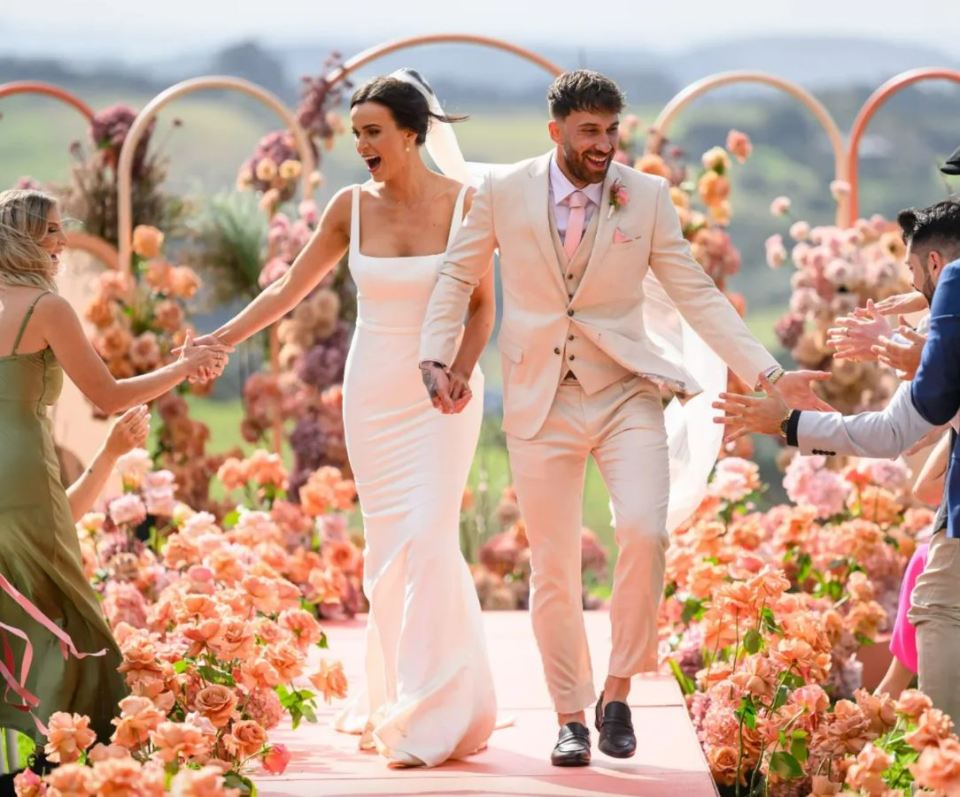 Billy Belcher and Sierah Swepstone from MAFS Australia hold hands and walk down an aisle covered in coral-colored flowers.