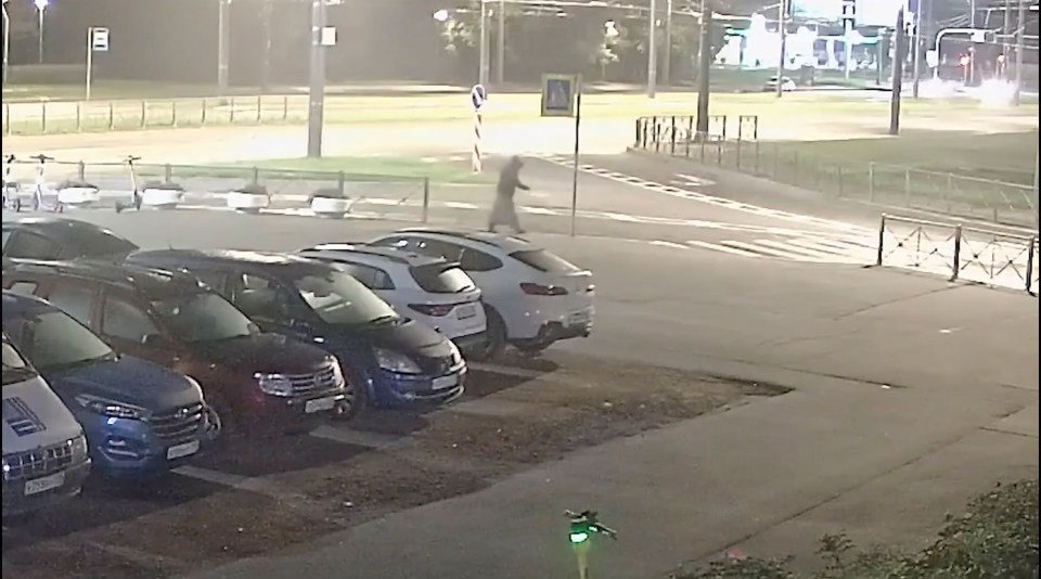 Security camera footage showing a person dressed in a light-colored dress walking past parked cars.