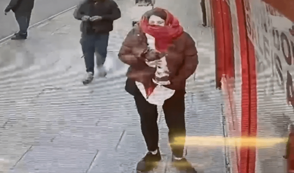 A person in a red scarf and brown jacket walking on a sidewalk.