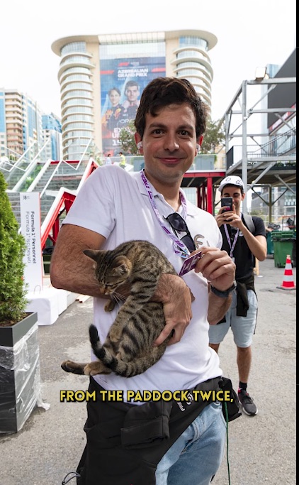 A man holding a striped cat with a F1 Grand Prix banner in the background.