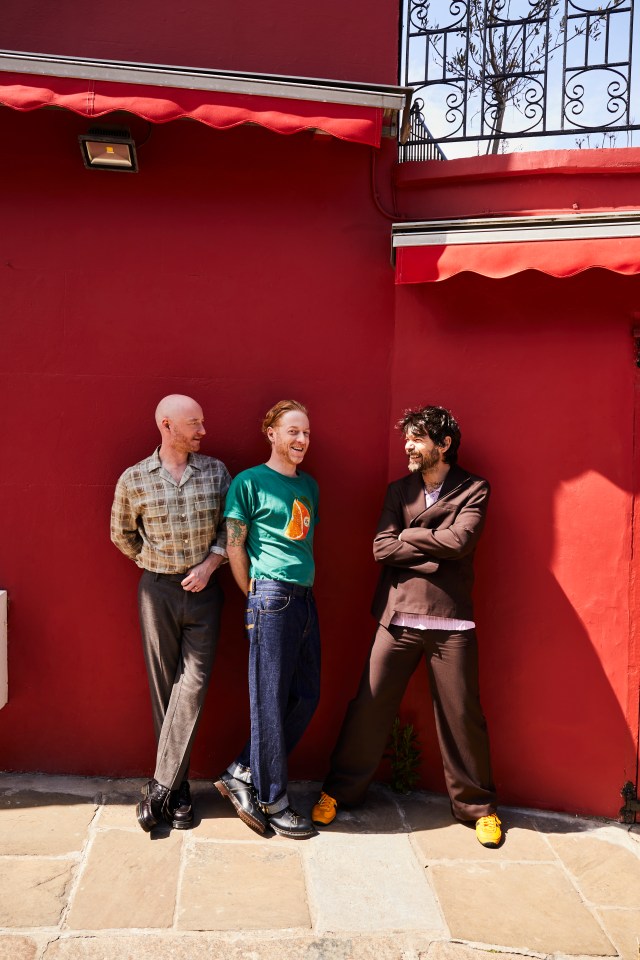 Biffy Clyro, three men, standing and laughing against a red wall with red awnings.