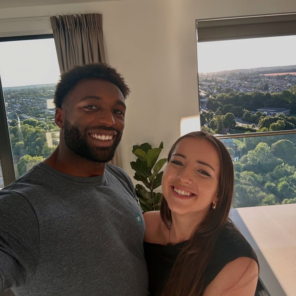 Paul Liba and his girlfriend smiling in front of a window with a city view.