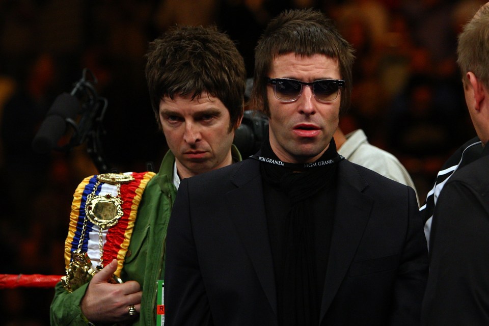 Noel Gallagher holding boxing belts and Liam Gallagher wearing sunglasses and a black suit at a boxing match.