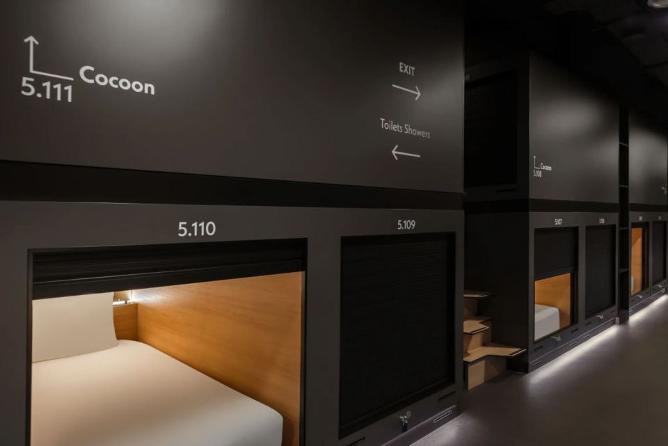 Interior of Zedwell Capsule Hotel, showing numbered capsule rooms with a sign pointing to "Exit" and "Toilets Showers".