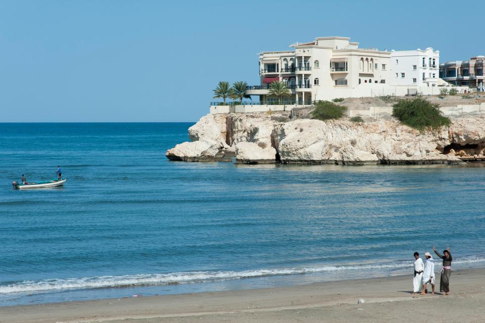 Qurum beach in Muscat, Oman, with a house on a cliff overlooking the sea, a boat with two men, and three men walking on the sand.
