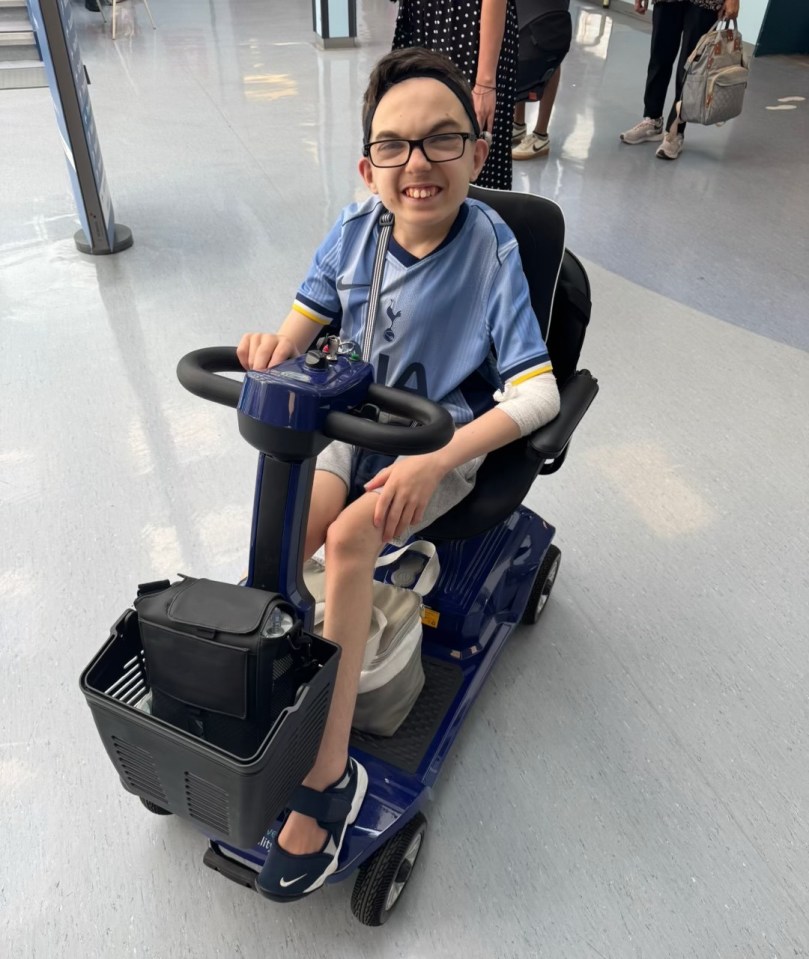 A boy in a light blue soccer jersey and glasses smiles while sitting in a dark blue mobility scooter.
