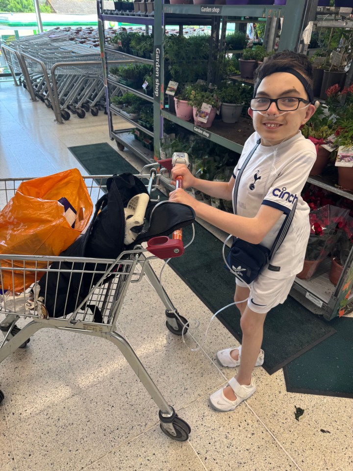 A young boy with nasal cannula tubing attached to a shopping cart smiles at the camera in a store.