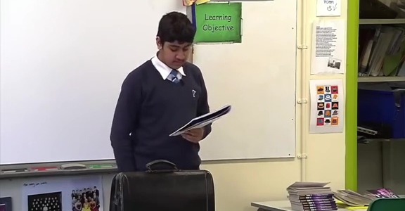 A male student wearing a navy blue uniform stands in a classroom, looking at papers in his hand.