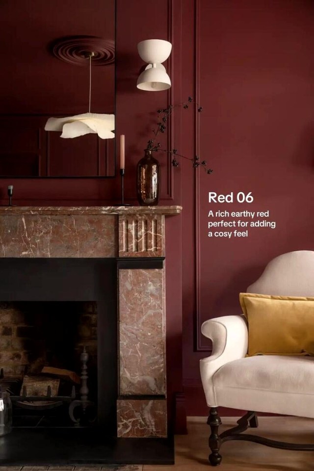 A living room painted in a rich earthy red, featuring a fireplace with a marble mantel and a cream-colored armchair with a yellow pillow.