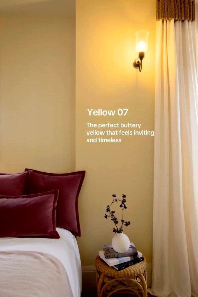 A bedroom painted in a buttery yellow color, with crimson pillows on the bed and white curtains, showcasing a specific interior design color trend for 2026.