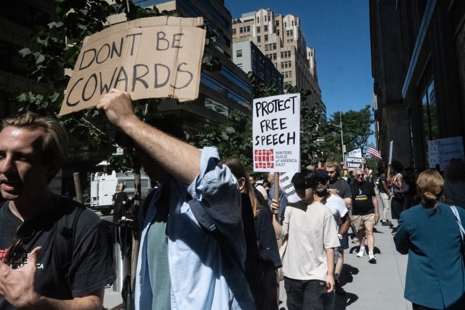 People protest holding signs that read "DON'T BE COWARDS" and "PROTECT FREE SPEECH" from the Writers Guild of America East.