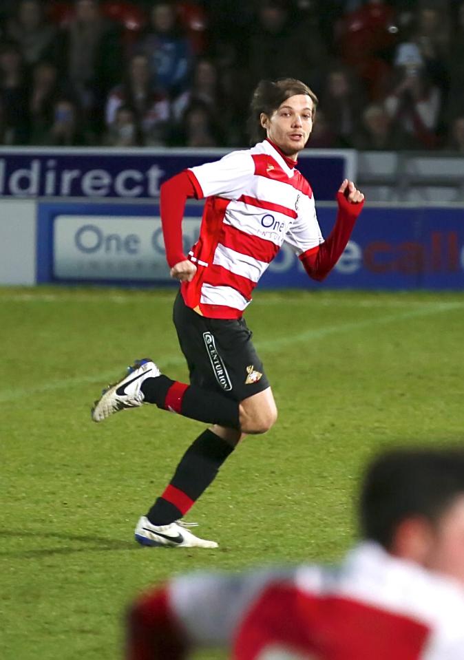 Louis Tomlinson playing football in a red and white striped jersey, black shorts, and white cleats with black socks.