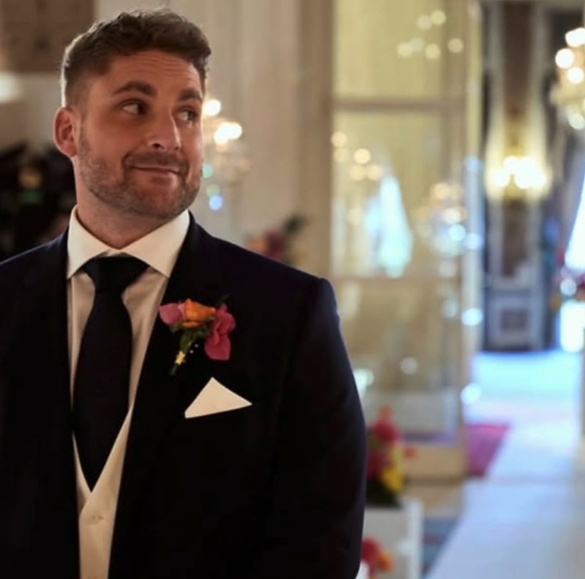 Ashley Dommett from Married at First Sight UK, in a suit with a boutonnière, looking sideways.