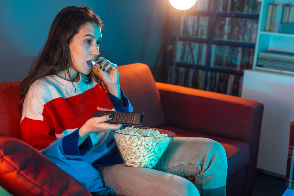 Woman watching TV and eating popcorn on a couch.