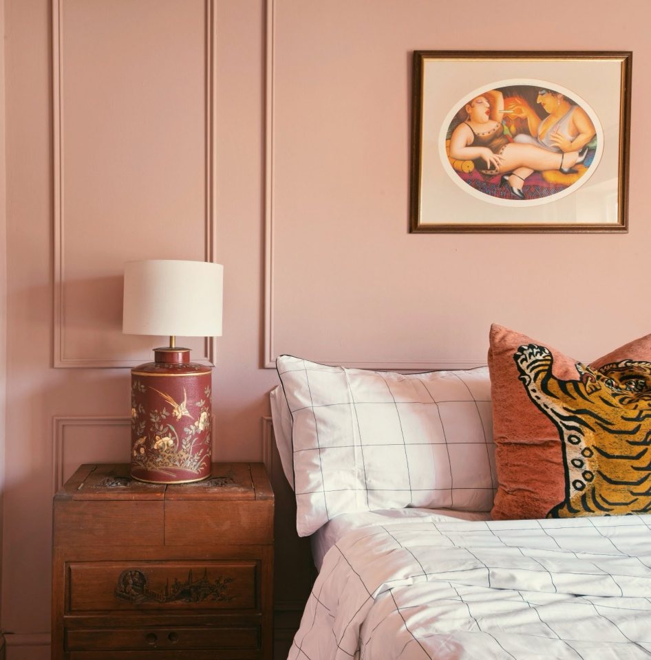 Bedroom with pink walls, a bed with gridded bedding and a tiger pillow, and a nightstand with a decorative lamp.