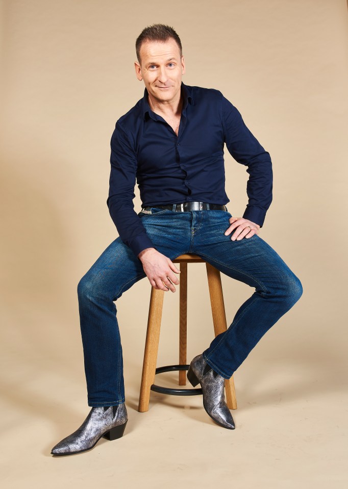 Nick Harding, 50, in a navy blue shirt and jeans, sitting on a stool wearing silver heeled boots.