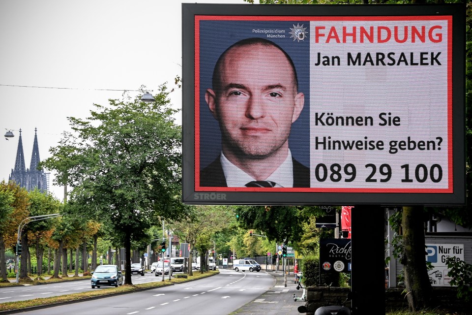 A digital wanted poster of Jan Marsalek displayed on an outdoor screen on a street in Cologne, Germany, with the Cologne Cathedral in the background.