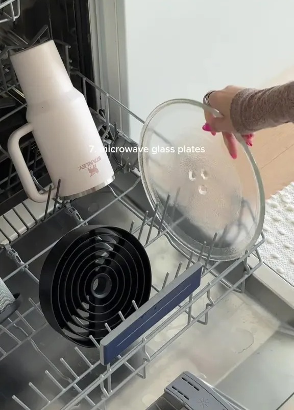 A person placing a microwave glass plate, a Stanley cup, and a spiral-shaped item into a dishwasher.