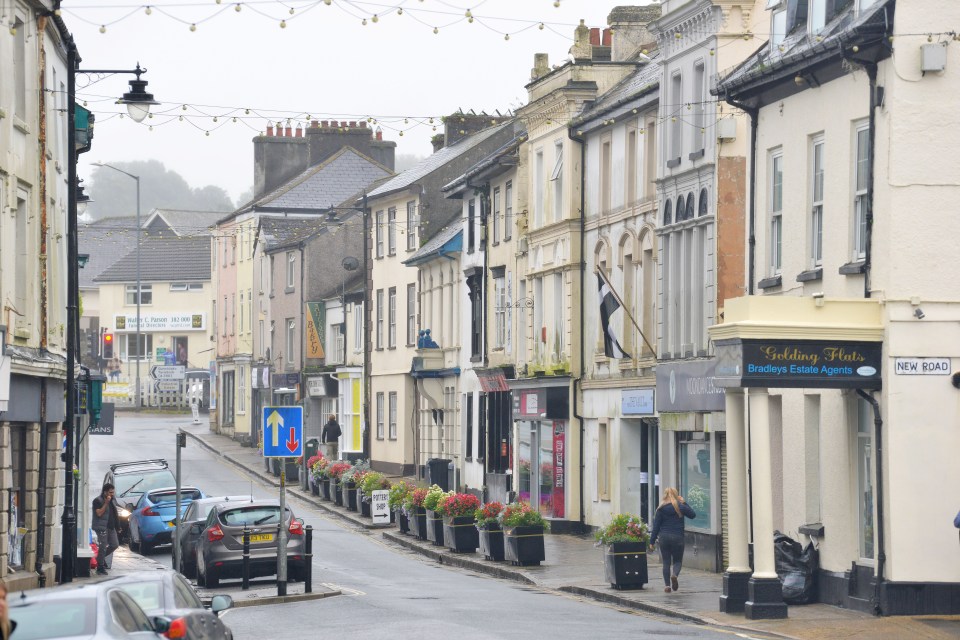 A street in Callington, Cornwall with cars parked along the side and a person walking on the sidewalk.