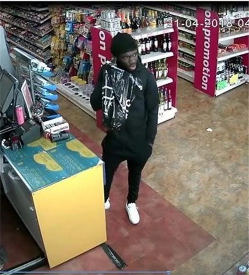 A man in a black hat, black hoodie, and black pants stands in a store holding a black plastic bag.