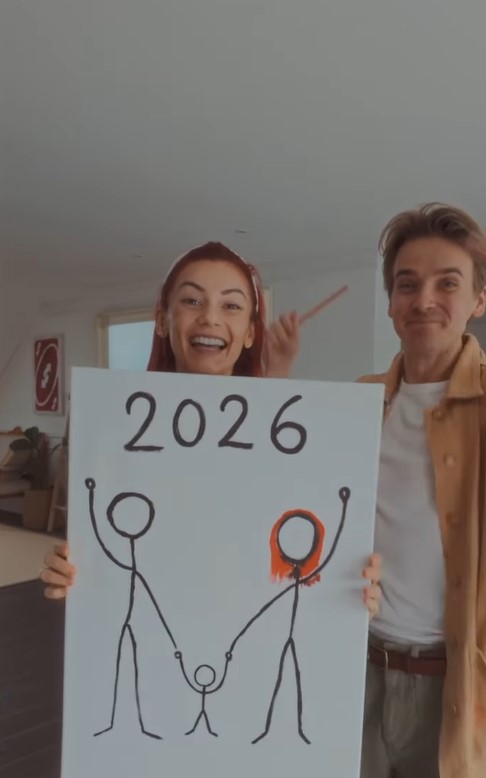 Couple holding a sign with a drawing of a family and "2026" written above.