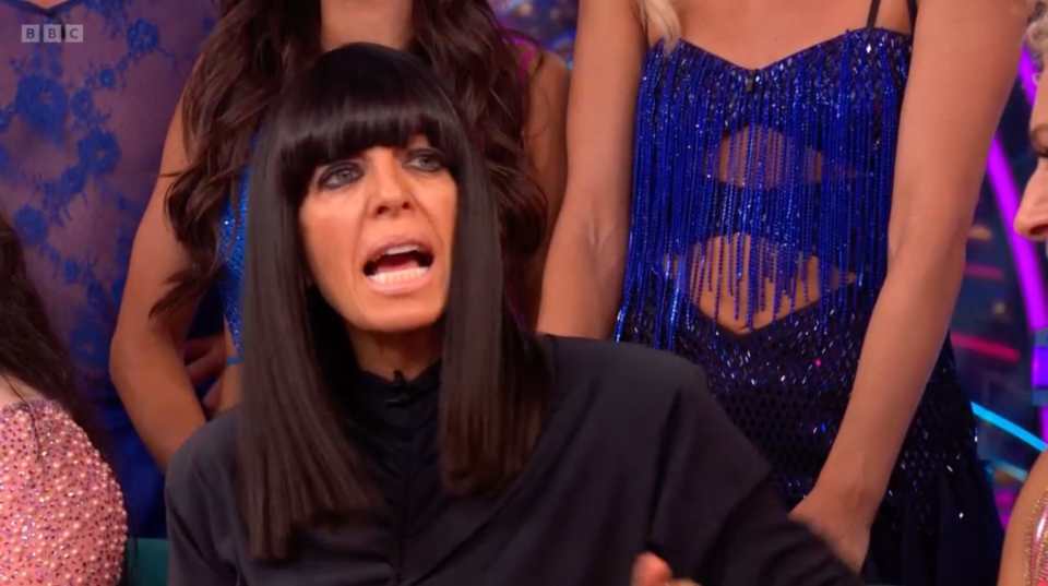 BBC presenter Claudia Winkleman speaking with her mouth open, surrounded by dancers in sparkly outfits.
