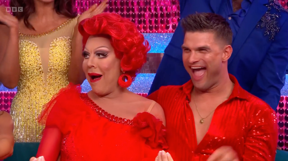 A drag queen in a red wig and a man in a red sequined shirt laughing.