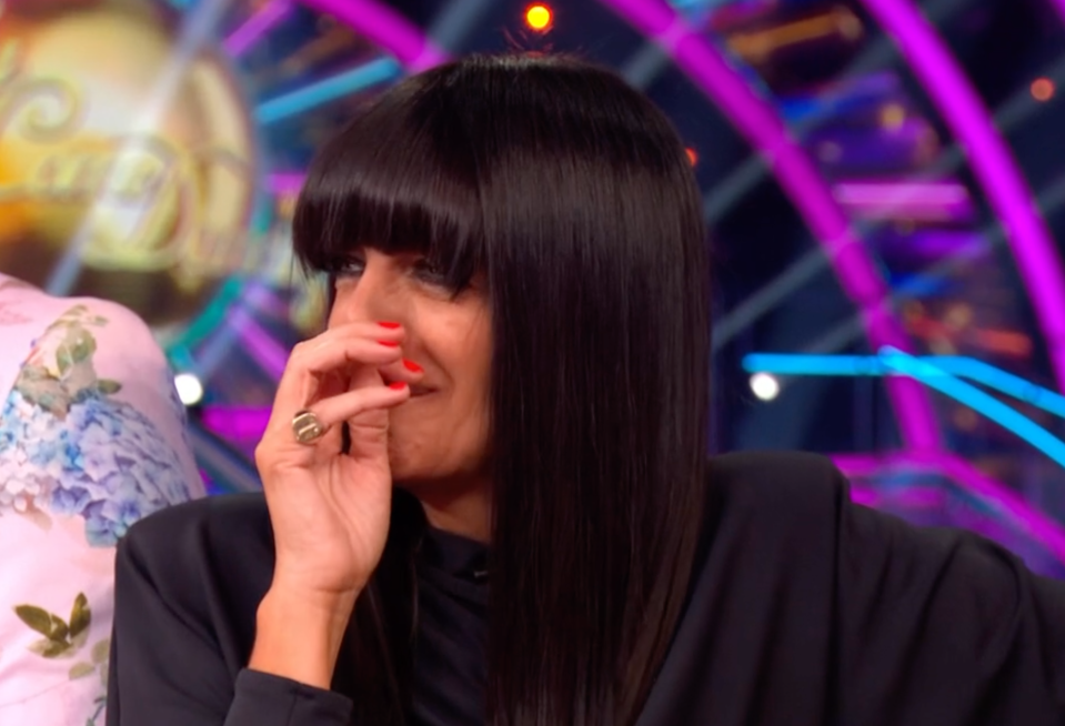 A woman with dark hair covering her forehead, wearing a black shirt, is seen from the chest up, covering her mouth with her hand.