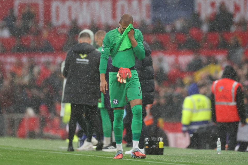 Robert Sanchez of Chelsea looks dejected, holding his face in a green towel during a rainy match.