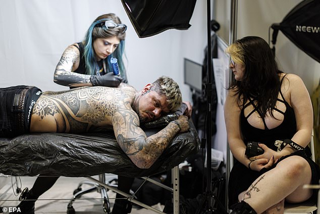 Another man, covered in tattoos, has another piece added to his back by an artist